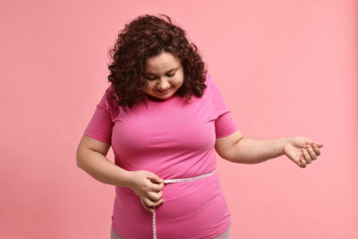 Weight loss. Happy plus size woman measuring waist with tape on pink background