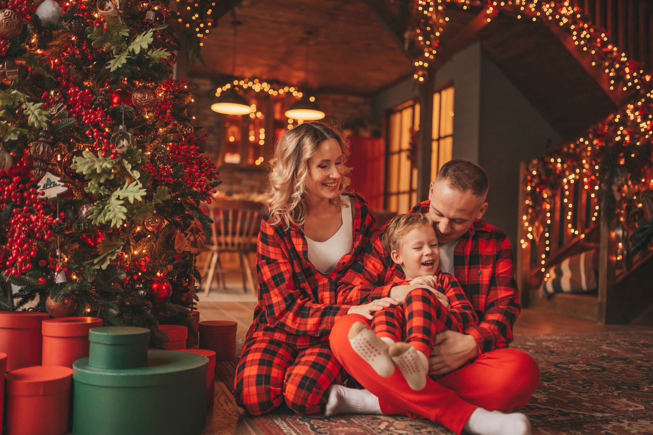 Smile active parents with small son in red checkered sleepwears waiting Santa indoor. Stylish family love laughing hugs kisses at home. Celebrate New year garlands and lights eve 25 December Noel tree