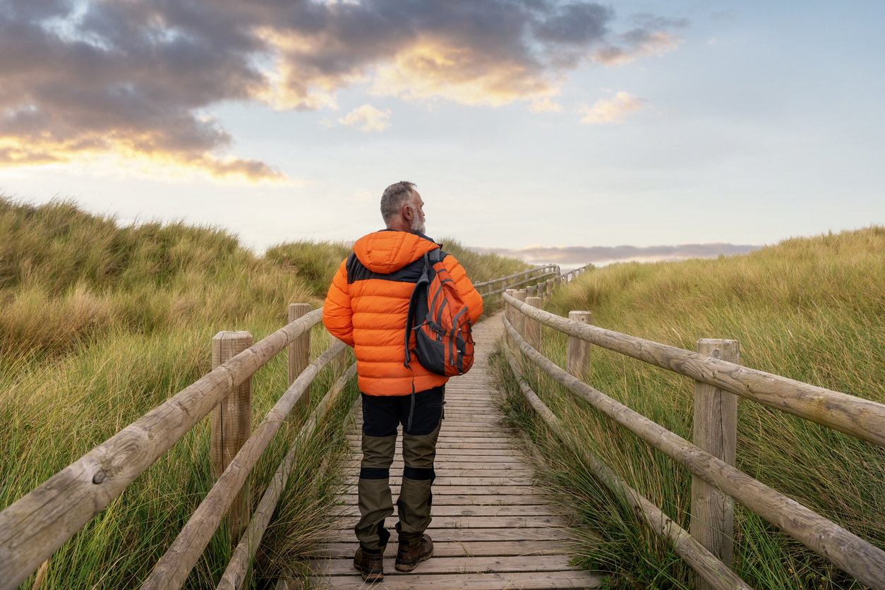 A man wearing an orange jacket strolls on a wooden boardwalk surrounded by tall grass, heading toward a picturesque sunset backdrop and enjoying nature's beauty.