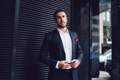 Confident smart businessman in dark suit with red scarf in pocket with mobile phone standing nearby metal wall and waiting looking away in downtown