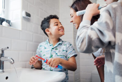 Children are brushing their teeth in the bathroom at home. The mother is checking the little boy's mouth to make sure he has brushed properly.