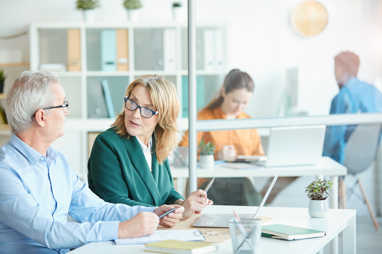 Mature businesswoman in eyeglasses talking to the mature businessman while they sitting at desk with laptop at office