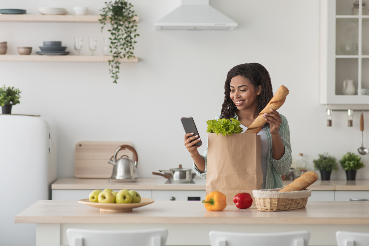Online buying food and grocery. Delivery service and mobile app. Happy young african american woman sort vegetables from eco package in minimalist kitchen interior, looks at phone, typing message