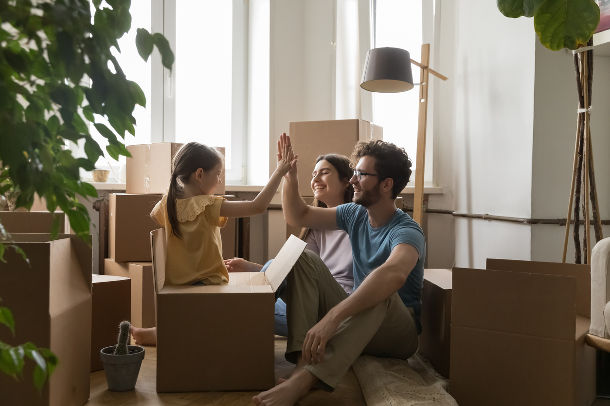 Young family with little daughter celebrate relocation day, feel happy, giving high five gesture seated on floor near heap of cardboard boxes.