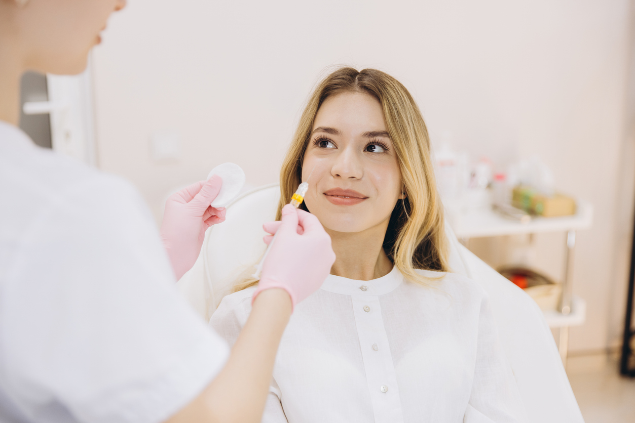 Cosmetologist wearing pink gloves preparing botox injection, holding syringe and cotton pad near smiling patient in white shirt