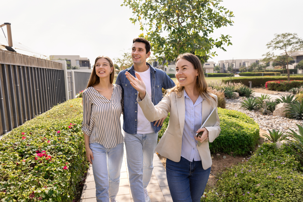 Happy professional real estate agent woman giving tour around well suburb neighborhood for house buyers, showing apartment exterior, location to young couple of customers, walking outside, pointing hand away