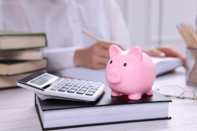 Piggy bank with calculator, book on white wooden table and woman working indoors, selective focus