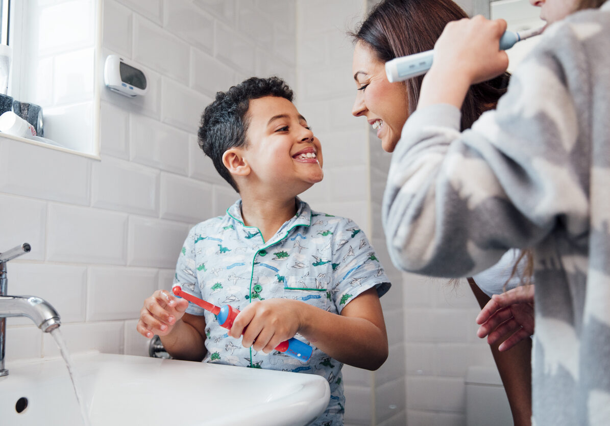 Children are brushing their teeth in the bathroom at home. The mother is checking the little boy's mouth to make sure he has brushed properly.