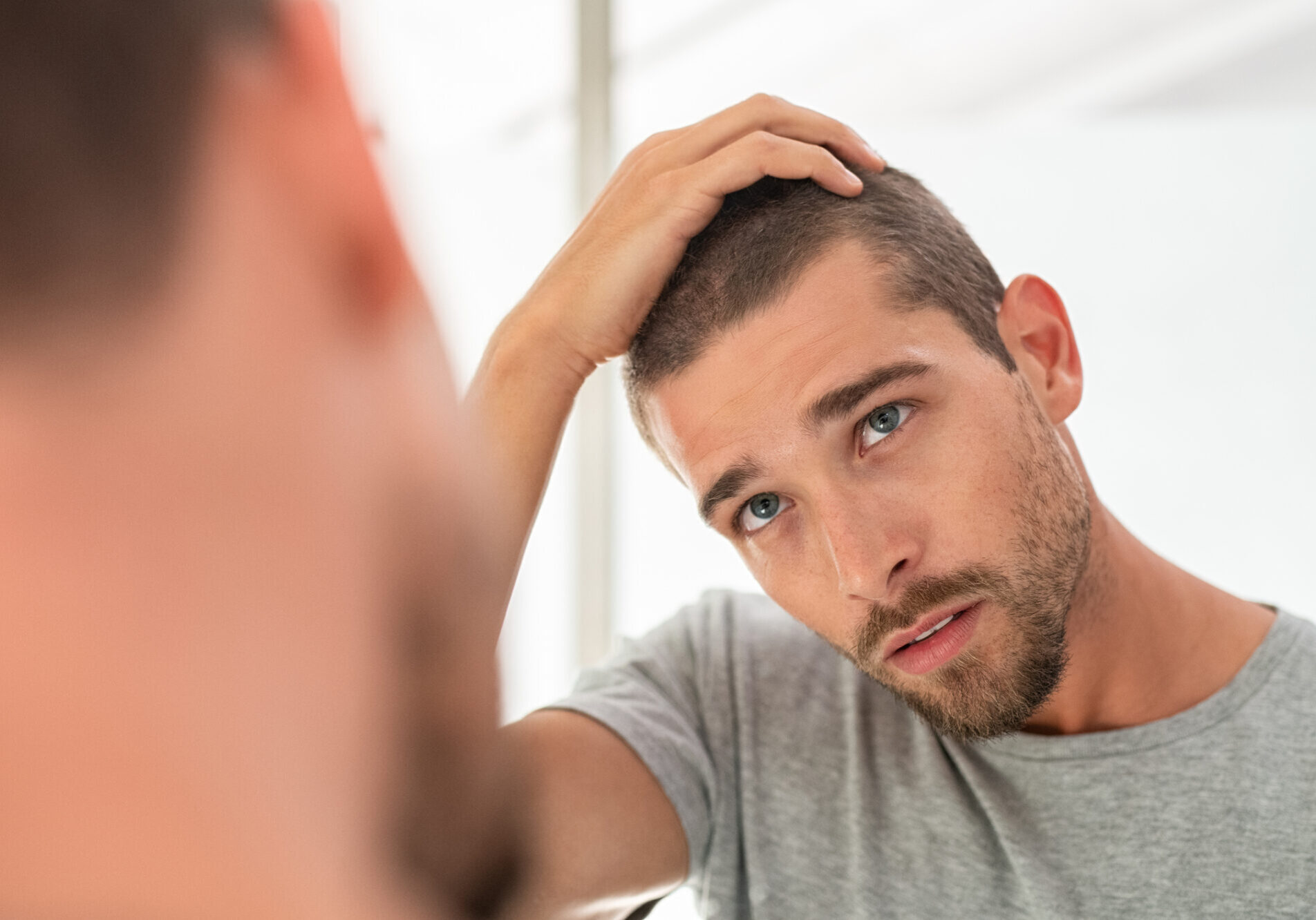 Young unshaven man looking at mirror in bathroom at home. Handsome guy looking at his face in mirror, checking hair and hairline. Man in pijamas concerned with hair loss.