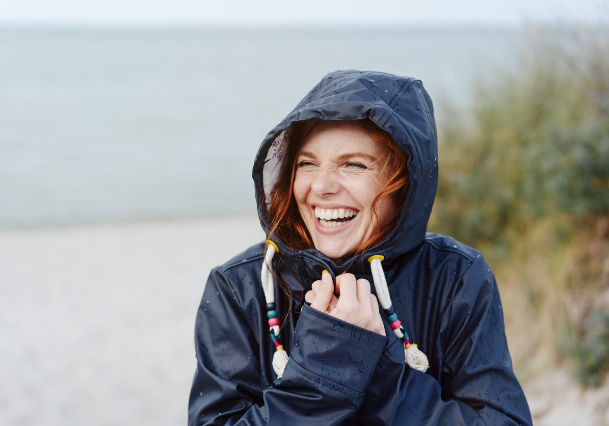 Laughing happy young woman embracing the cold autumn weather snuggling into her warm anorak with a beaming vivacious smile as she strolls along a beach