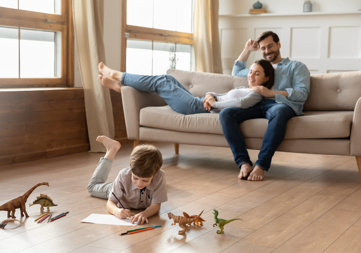 Smiling loving young bonding couple parents relaxing on comfortable sofa, watching little kid son drawing pictures in album, lying on wooden floor in living room, happy family weekend pastime.