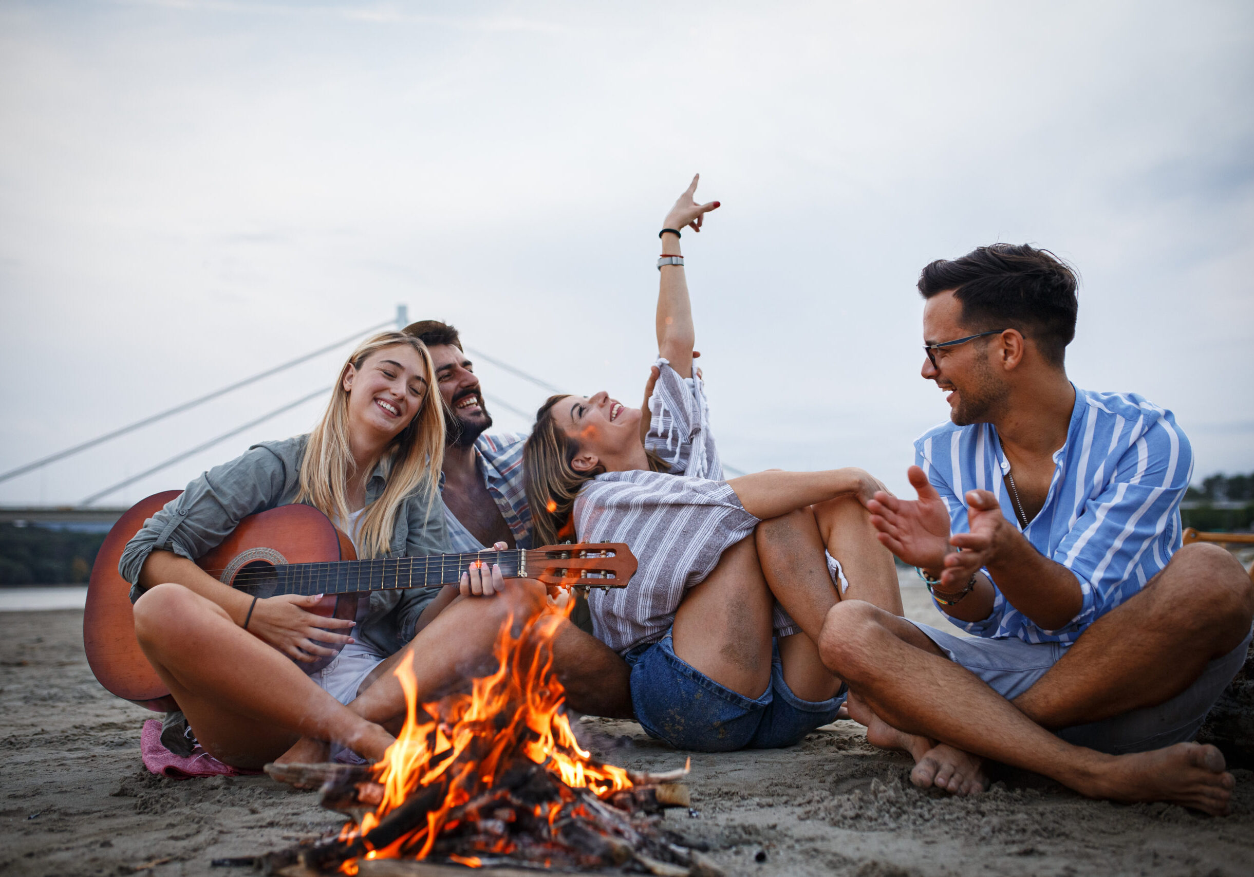 Group of friends sitting around camp fire at the beach at the autumn evening. They play guitar and singing.