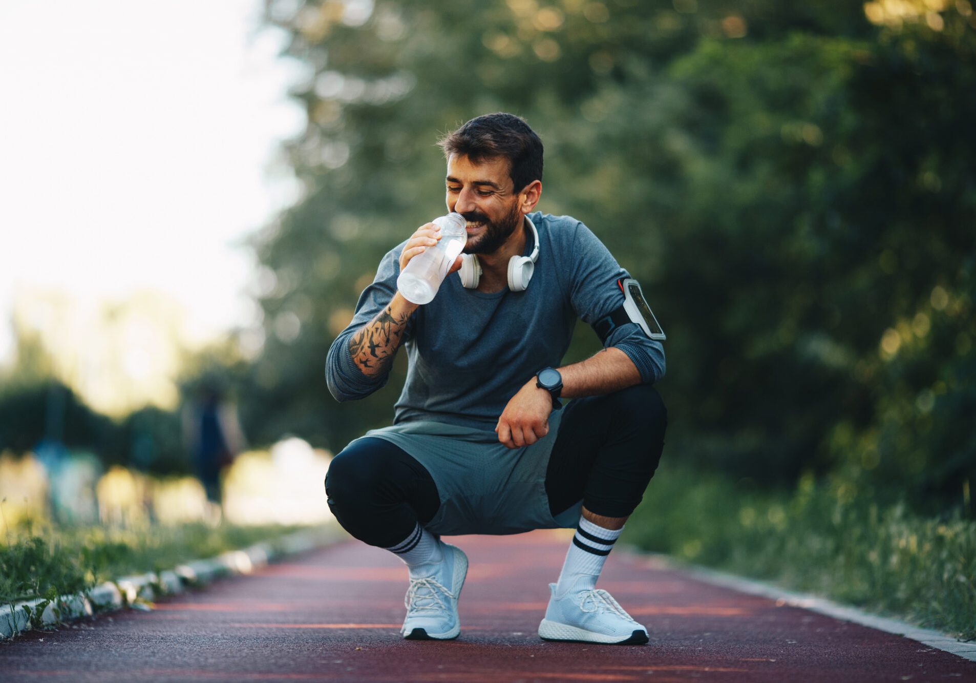 Young attractive smiling sportsman crouching on treadmill and drinking water.