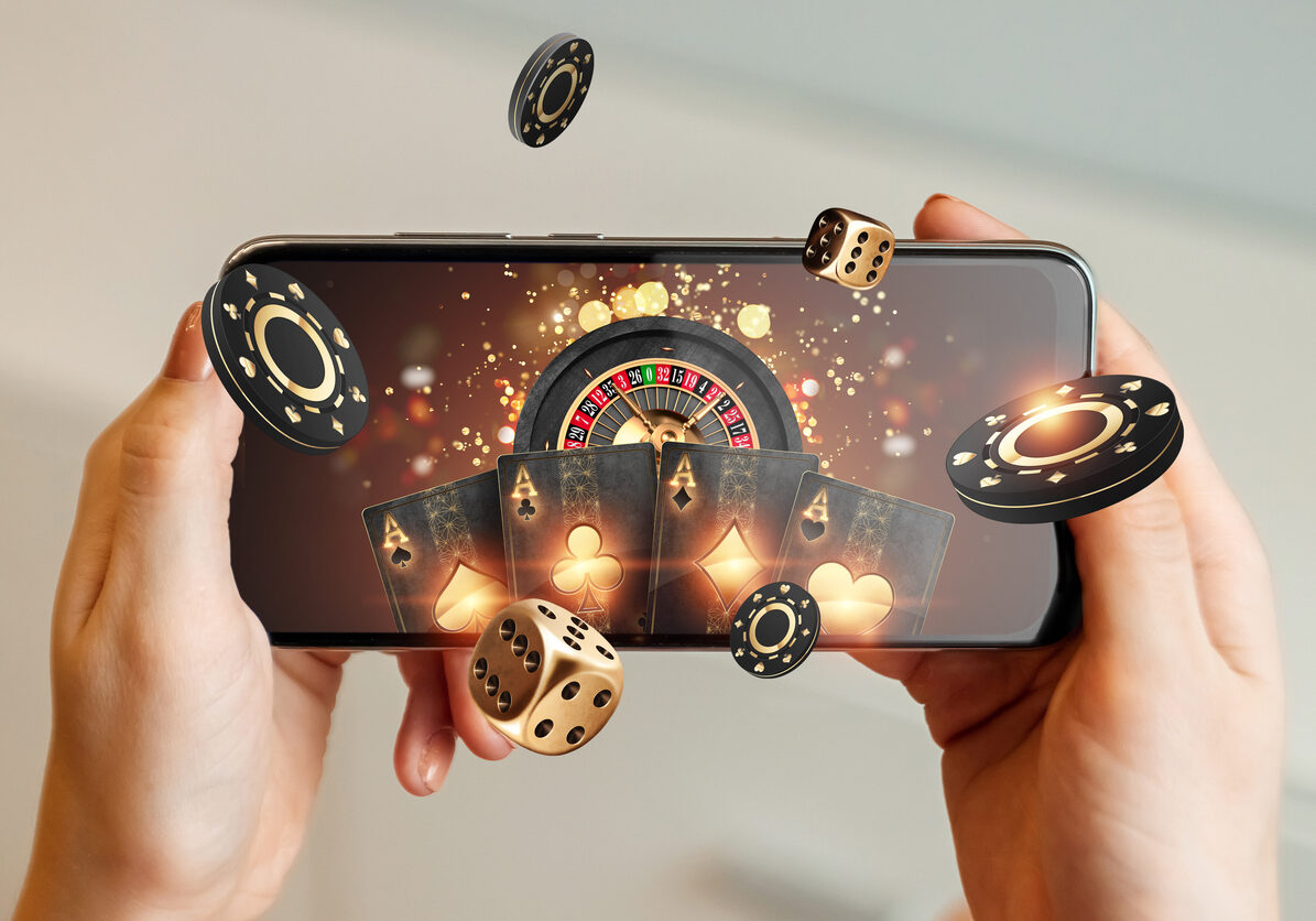 Creative background, online casino, in a man's hand a smartphone with playing cards, roulette and chips, black-gold background. Internet gambling concept. Copy space.