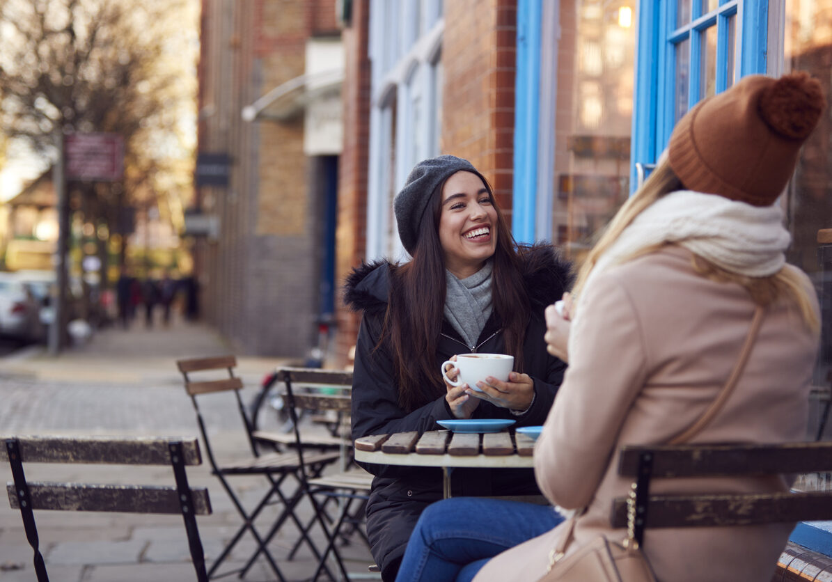 Two Female Friends Meeting Sitting Outside Coffee Shop On City High Street