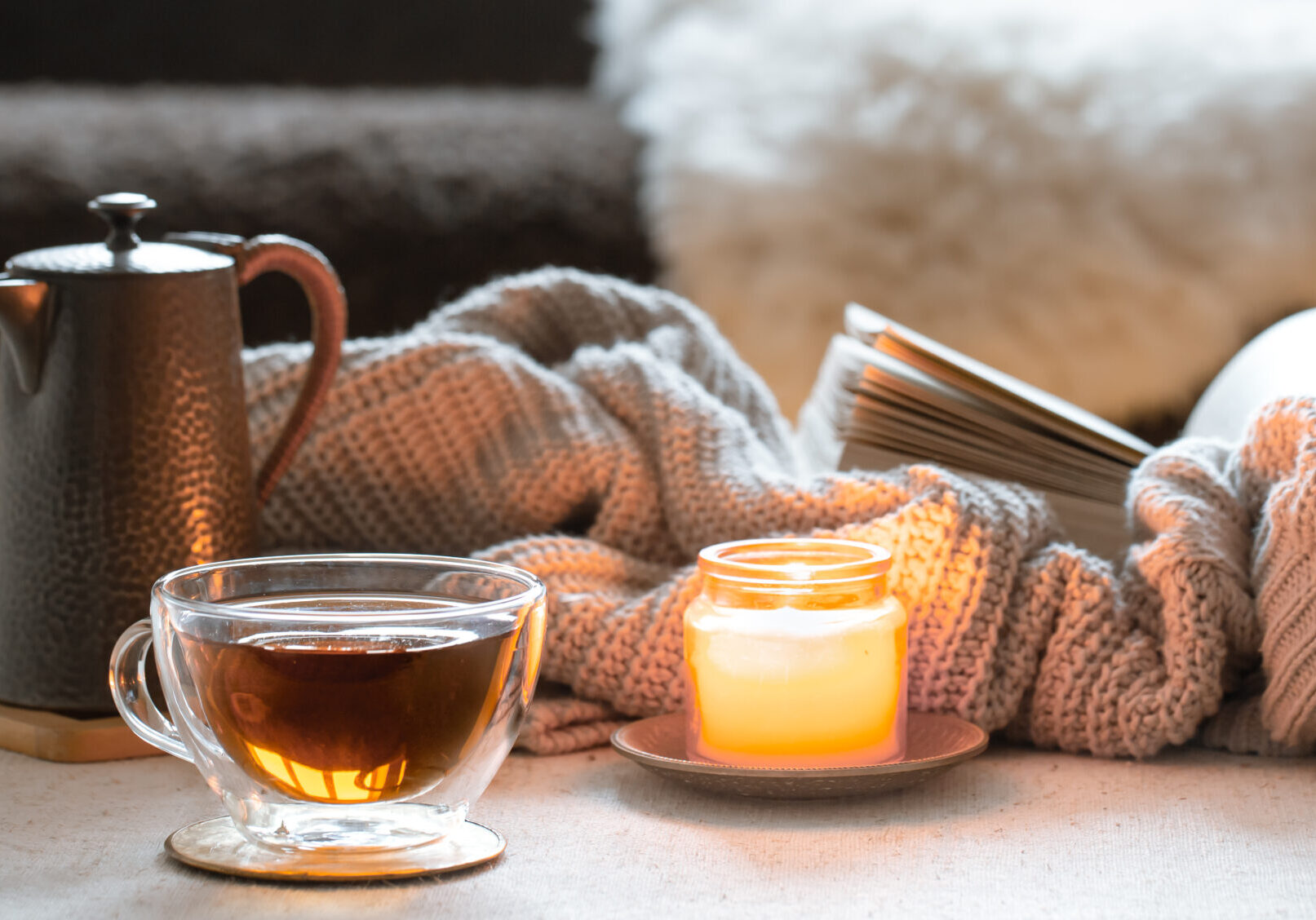 Glass cup of tea, teapot, candle and book with knitted element. The concept of home comfort and warmth.