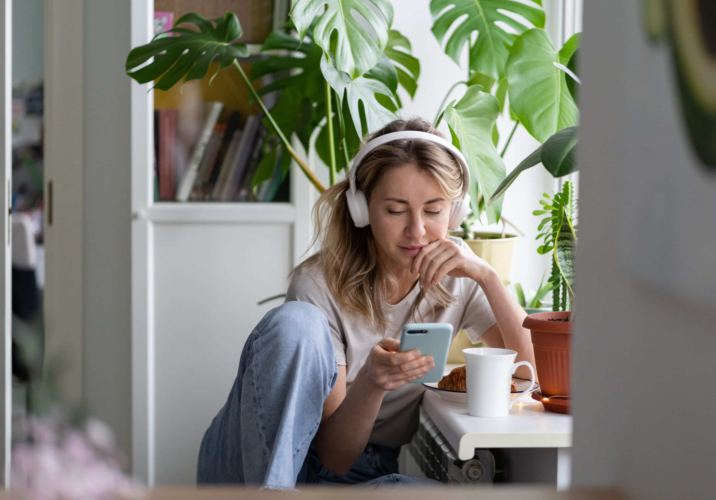 Woman listening to music, wear wireless white headphones, using mobile smart phone, chatting in social networks, sitting next to the window, houseplants on windowsill. Life at home. Time to relax.