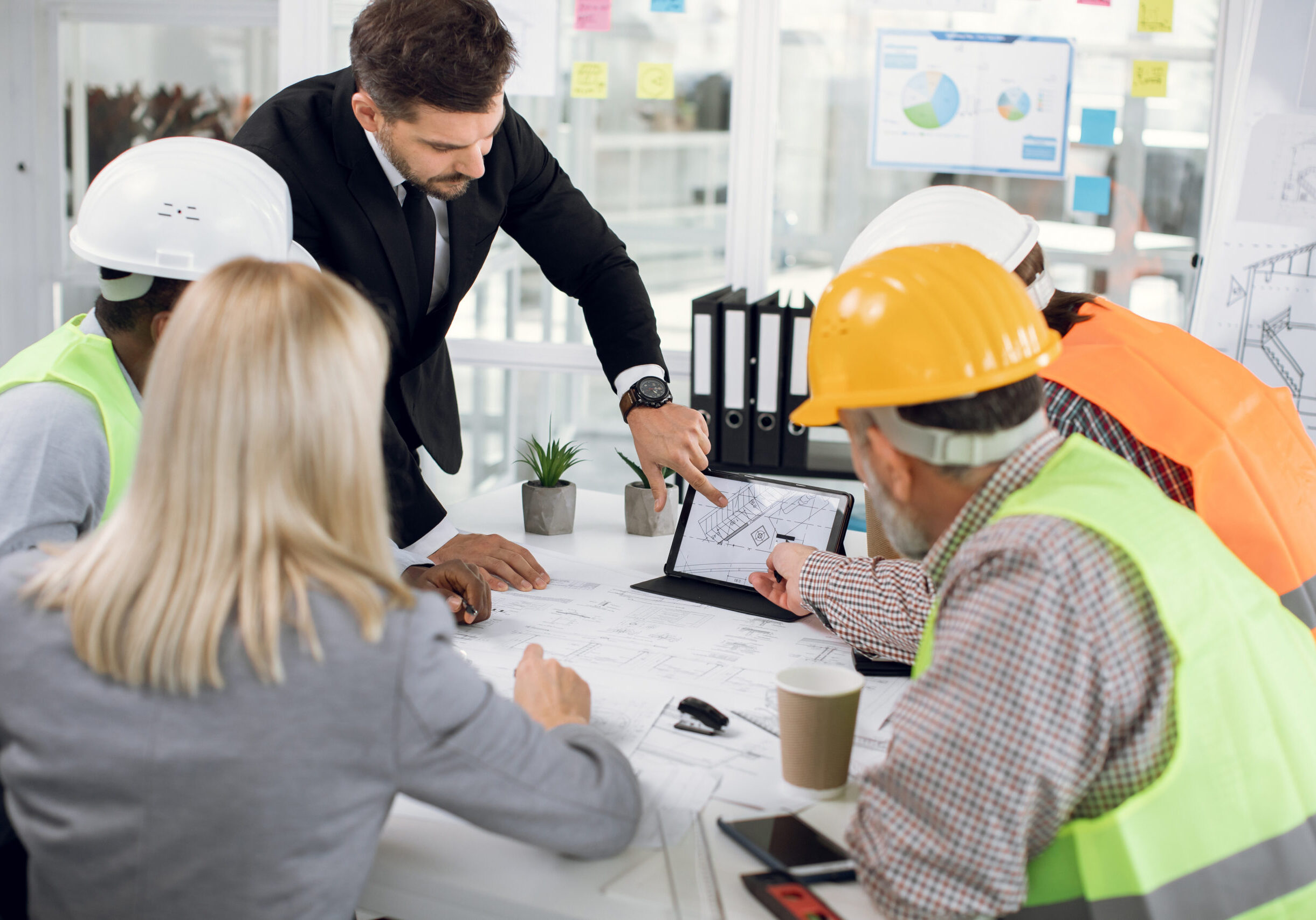 High-skilled purposeful multiracial team of engineers in special uniforms and male and female managers, brainstorming together over joint building project, looking at the tablet screen