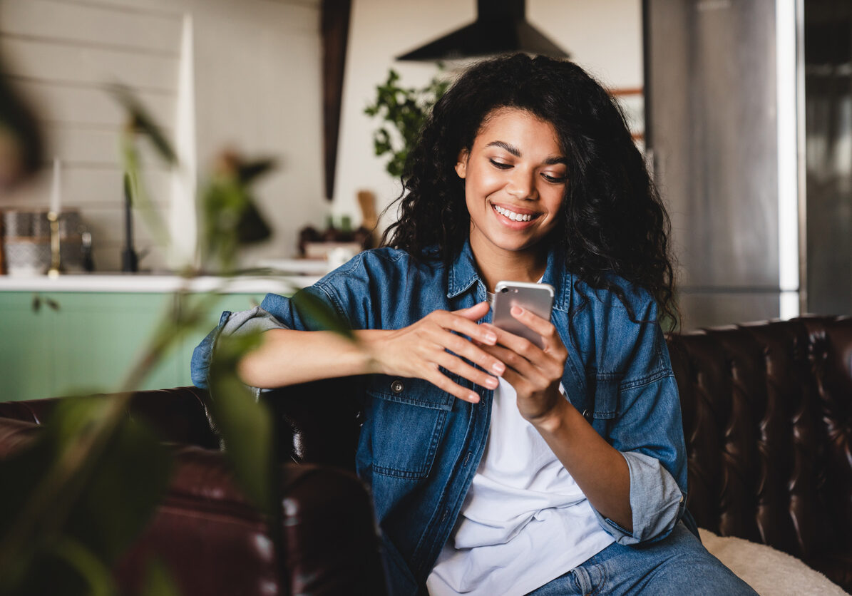 Relaxed young african woman texting on her phone in the modern house.Smiling african american woman using smartphone at home, messaging or browsing social networks while relaxing on couch