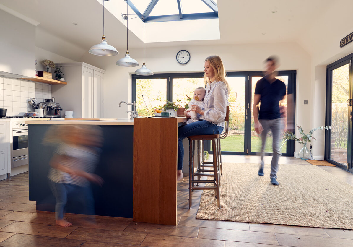 Busy Family Kitchen With Mother Working On Laptop And Holding Baby As Daughter Runs In Foreground