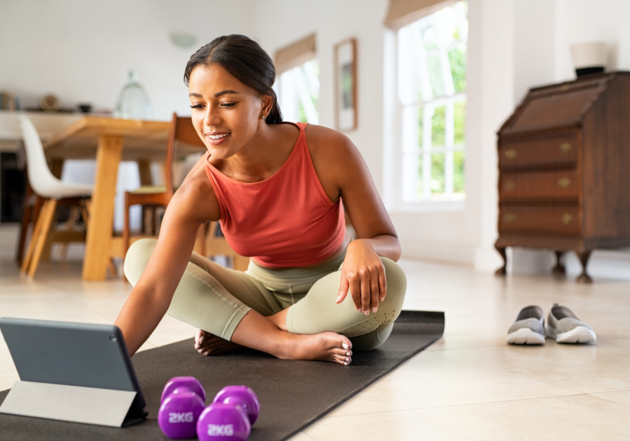 Mature healthy woman using app on digital tablet at home to train in hata yoga. Mature african american fit woman using digital tablet while sitting on yoga mat after fitness exercises. Happy smiling indian lady watching exercise tutorials on digital tablet while working out with dumbbells at home.