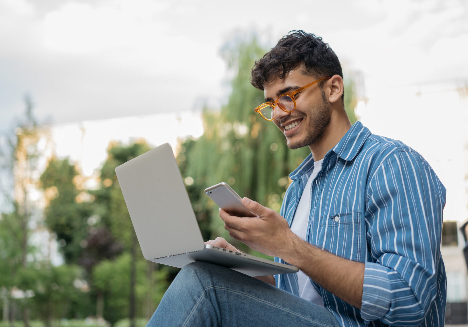 Young handsome Indian man using laptop computer, mobile phone, working freelance project online, sitting outdoors. Successful business. Asian student studying, learning language, online education concept