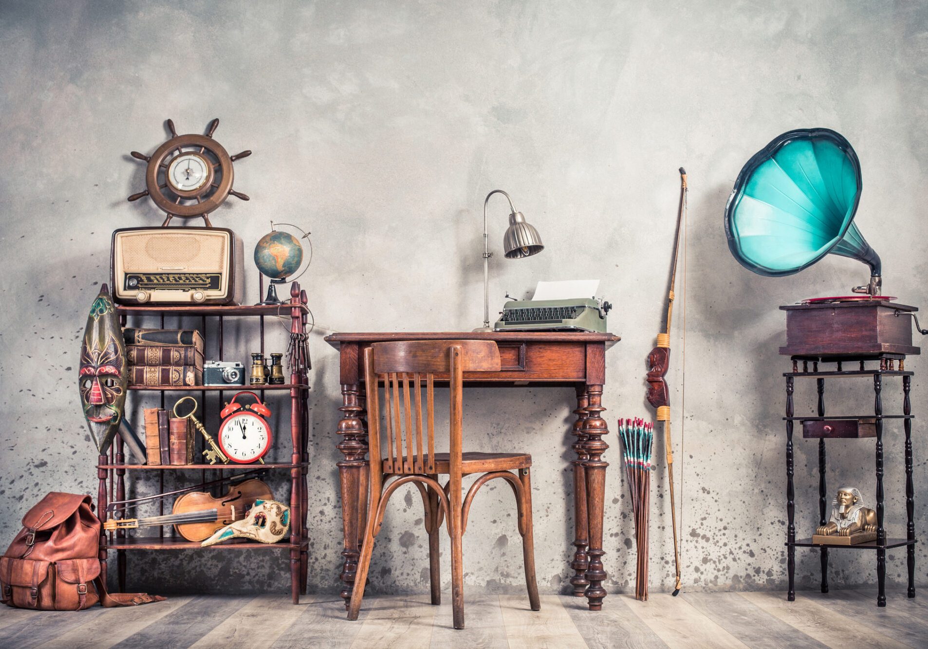 Typewriter on table, vintage gramophone, old books, radio, globe, binoculars, carnival mask, camera, fiddle on shelf, steering wheel, plane, travel backpack, bow. Retro style filtered photo