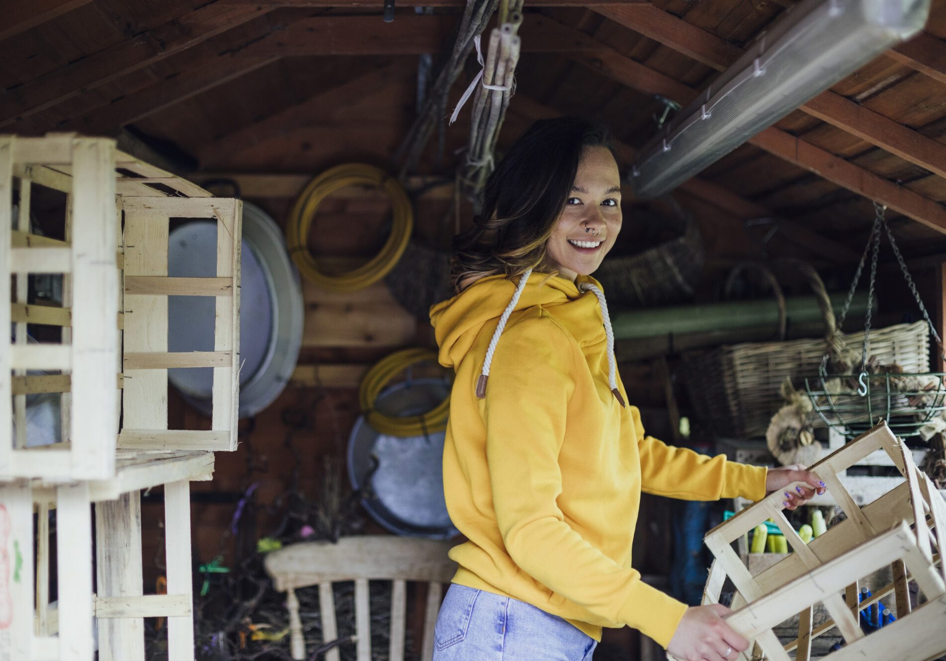A young woman standing in a shed looking at the camera and holding a wooden crate. She is organising the shed.
