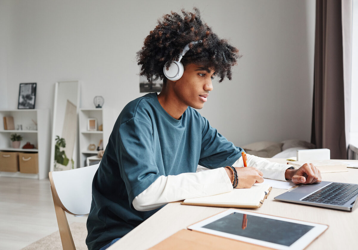 Side view portrait of African-American teenage boy studying at home or in college dorm and using laptop, copy space