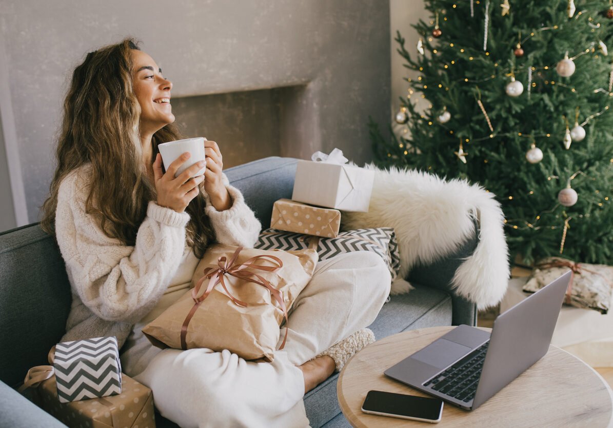 Young woman drinking hot coffee or cocoa sitting on couch in cozy room with Christmas tree. Winter holiday mood.