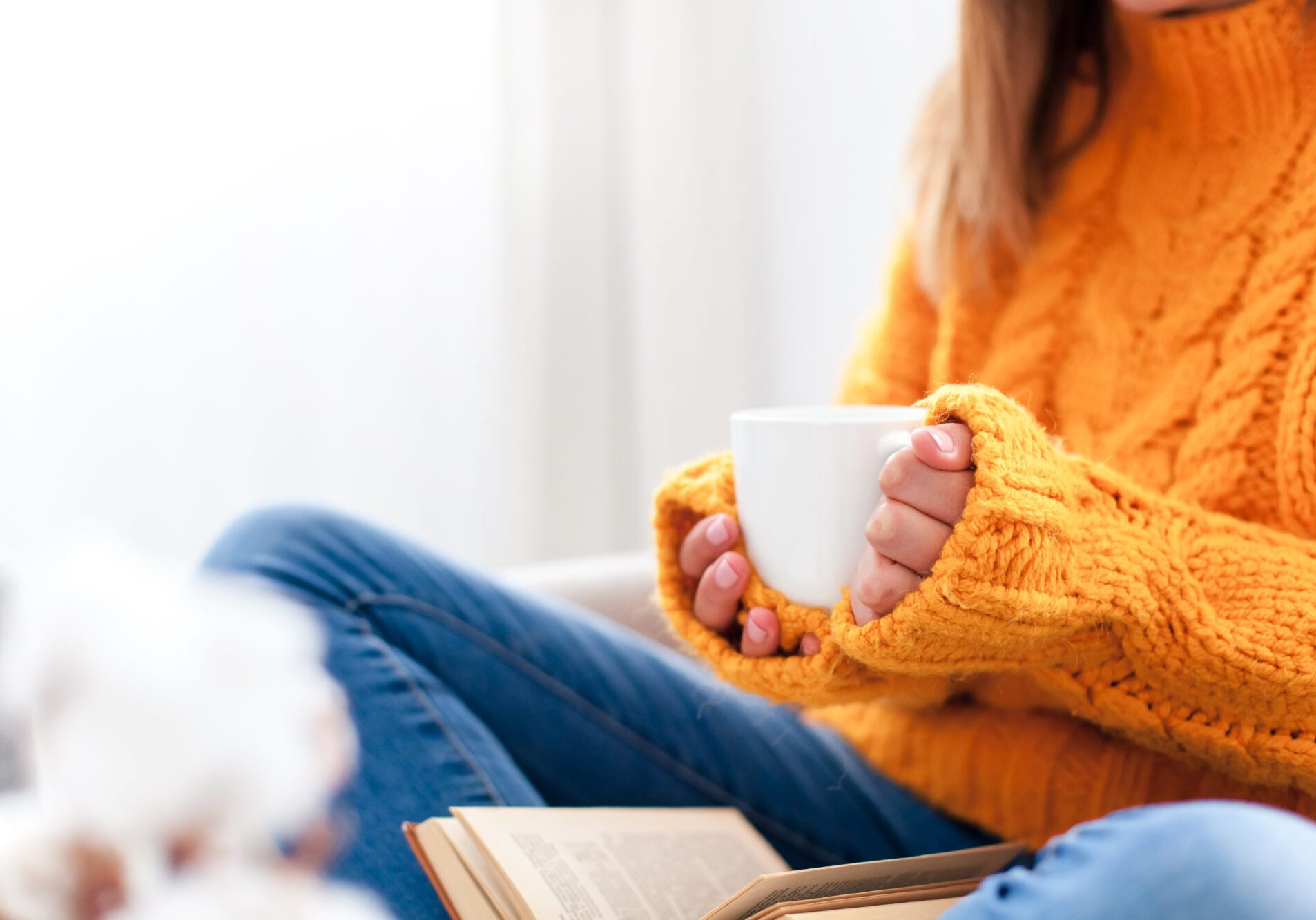 Young woman drinking coffee at home. Happy girl relaxing, reading book and enjoying cup of tea in cozy living room. Concept of digital detox, slow living, autumn moments. Close up of hands with mug.