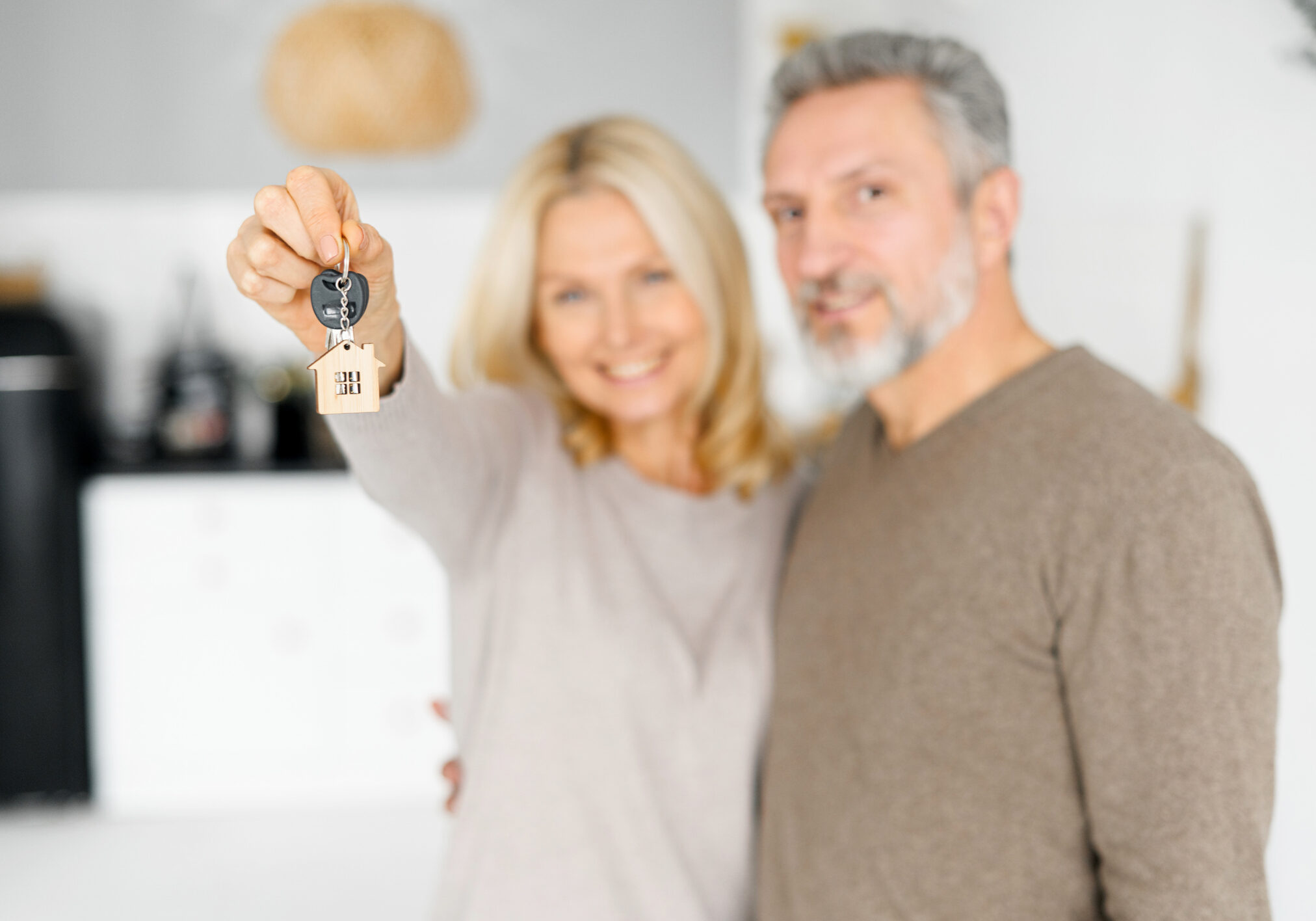 Selective focus on the keys with keychain in form of little house in female hand, happy cheerful middle-aged couple holds keys from new property, happy buyers of an own estate. Spouses get a mortgage