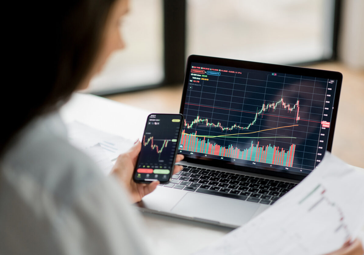 Successful smart woman investor and crypto trader, using laptop and smartphone, analyzes charts of trading in stock market and digital cryptocurrency exchange, conducts analysis, trading crypto coins