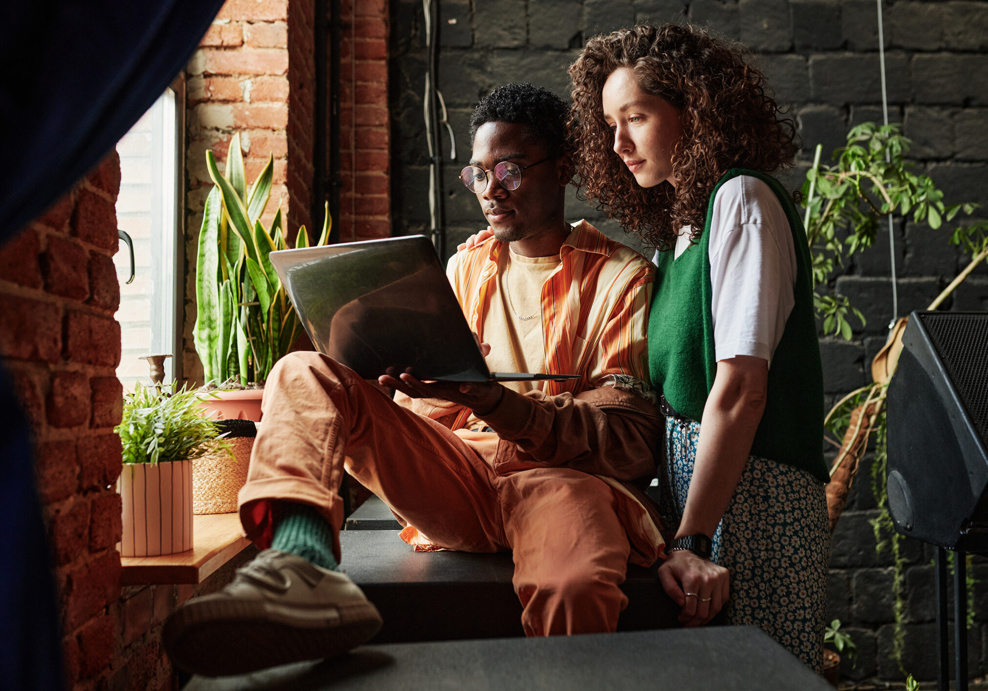 Young interracial couple working in the net while man holding laptop and his girlfriend standing next to him in front of window in studio