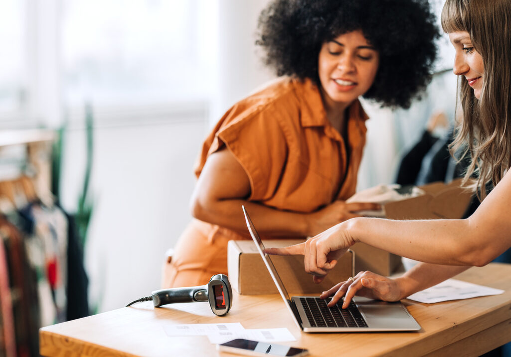 Clothing store owners having a discussion while preparing online orders for shipping. Two young women using a laptop together in a thrift store. Female entrepreneurs running an e-commerce small business.