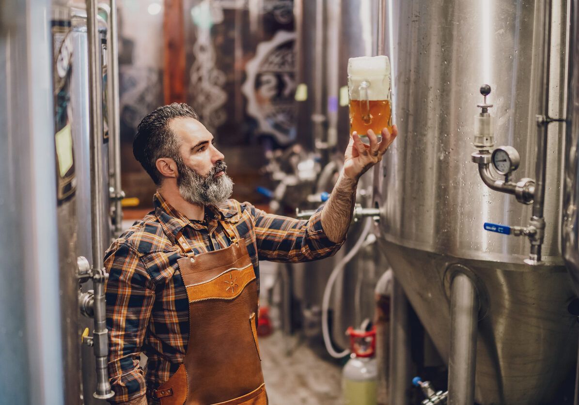 Bearded brewery master holding glass of beer and evaluating its visual characteristics. Small family business, production of craft beer.