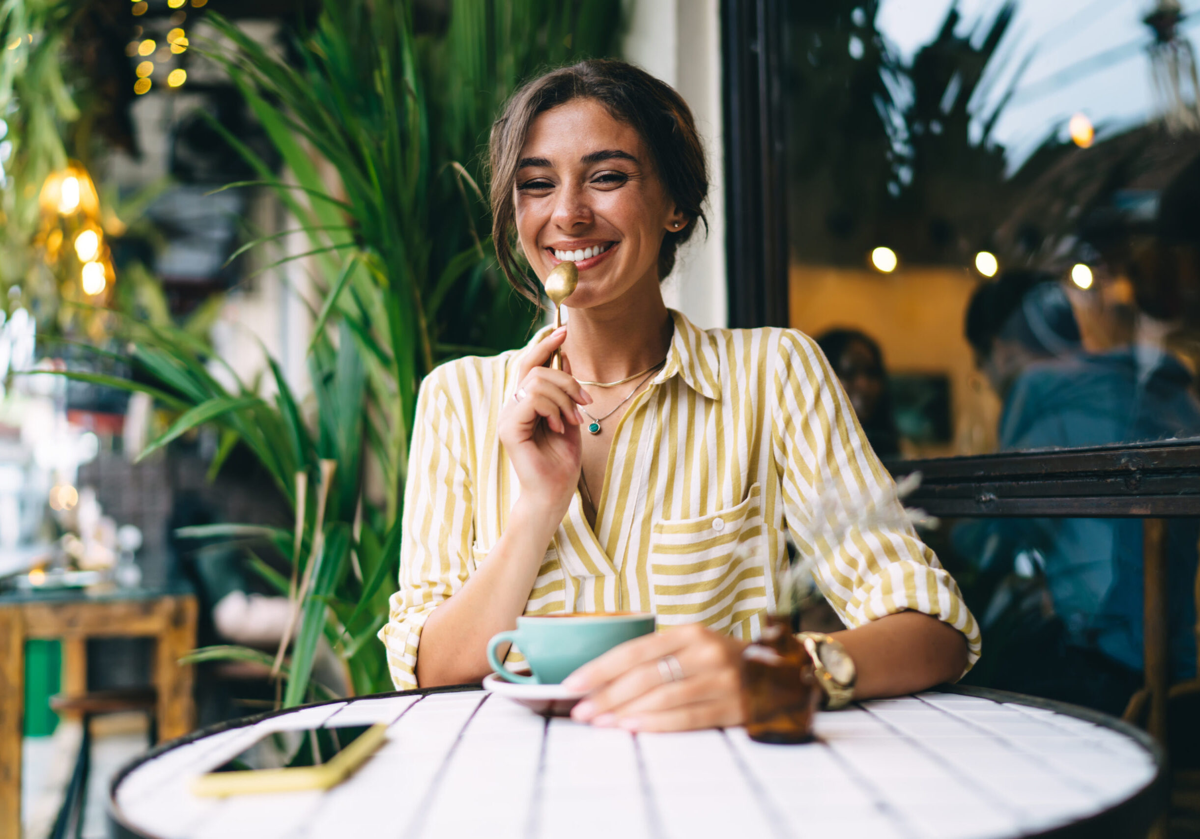 Optimistic young female wearing striped shirt drinking cup of fresh coffee and holding teaspoon while sitting at round table in open terrace
