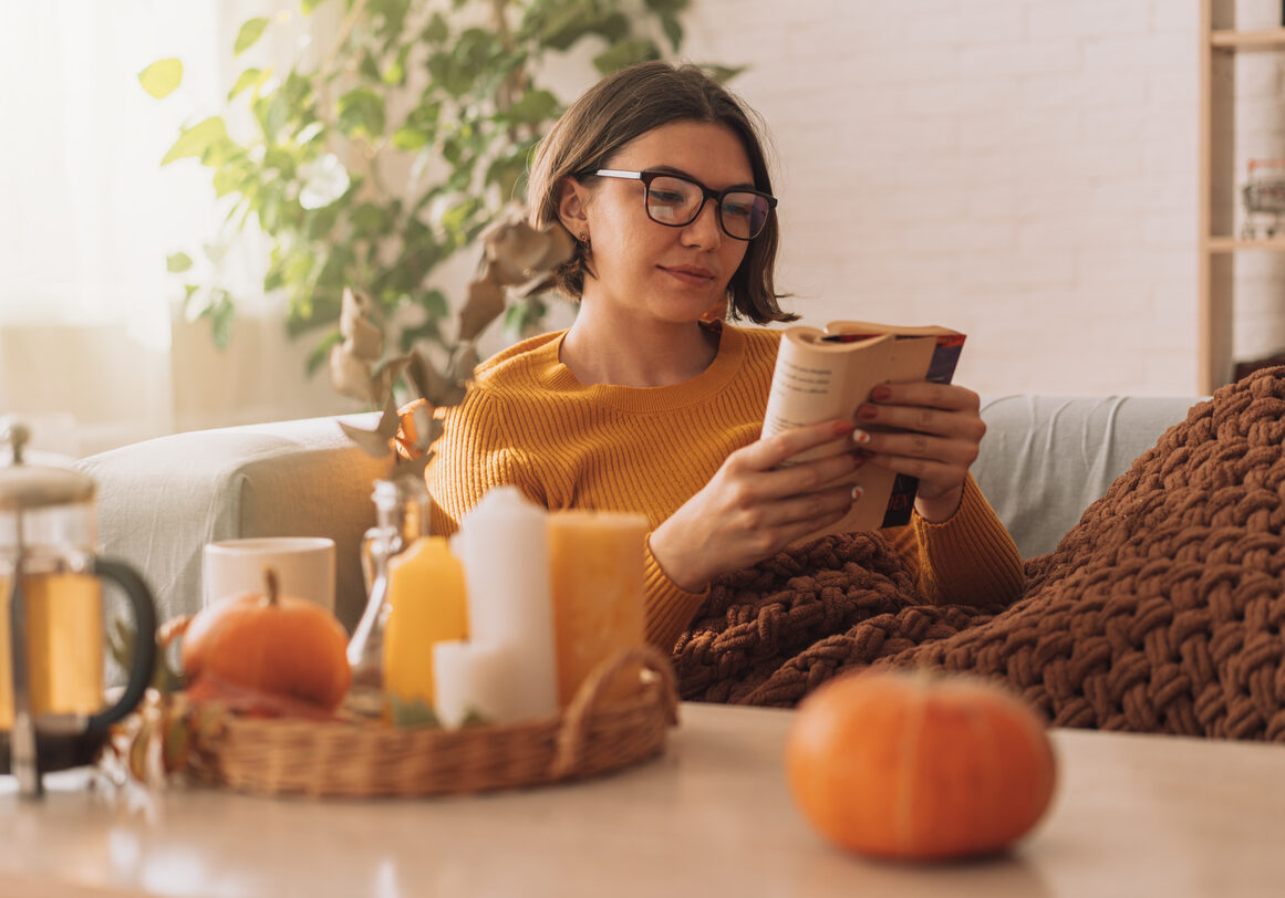 Lady reads a book in blanket on the sofa in front of teapot and candles.