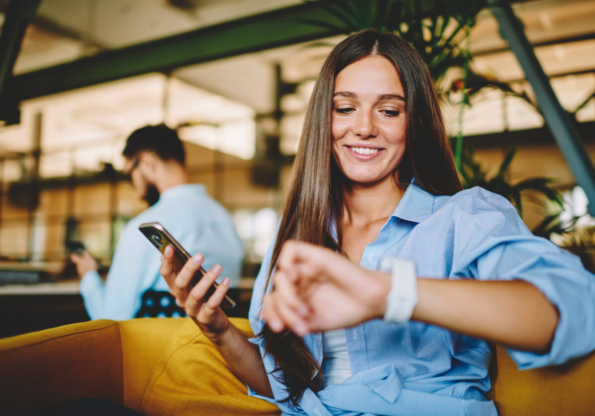 Carefree Caucasian woman 20s checking time on wearable smartwatch during leisure recreation with digital tablet, casual dressed millennial female using modern technology and wrist accessory