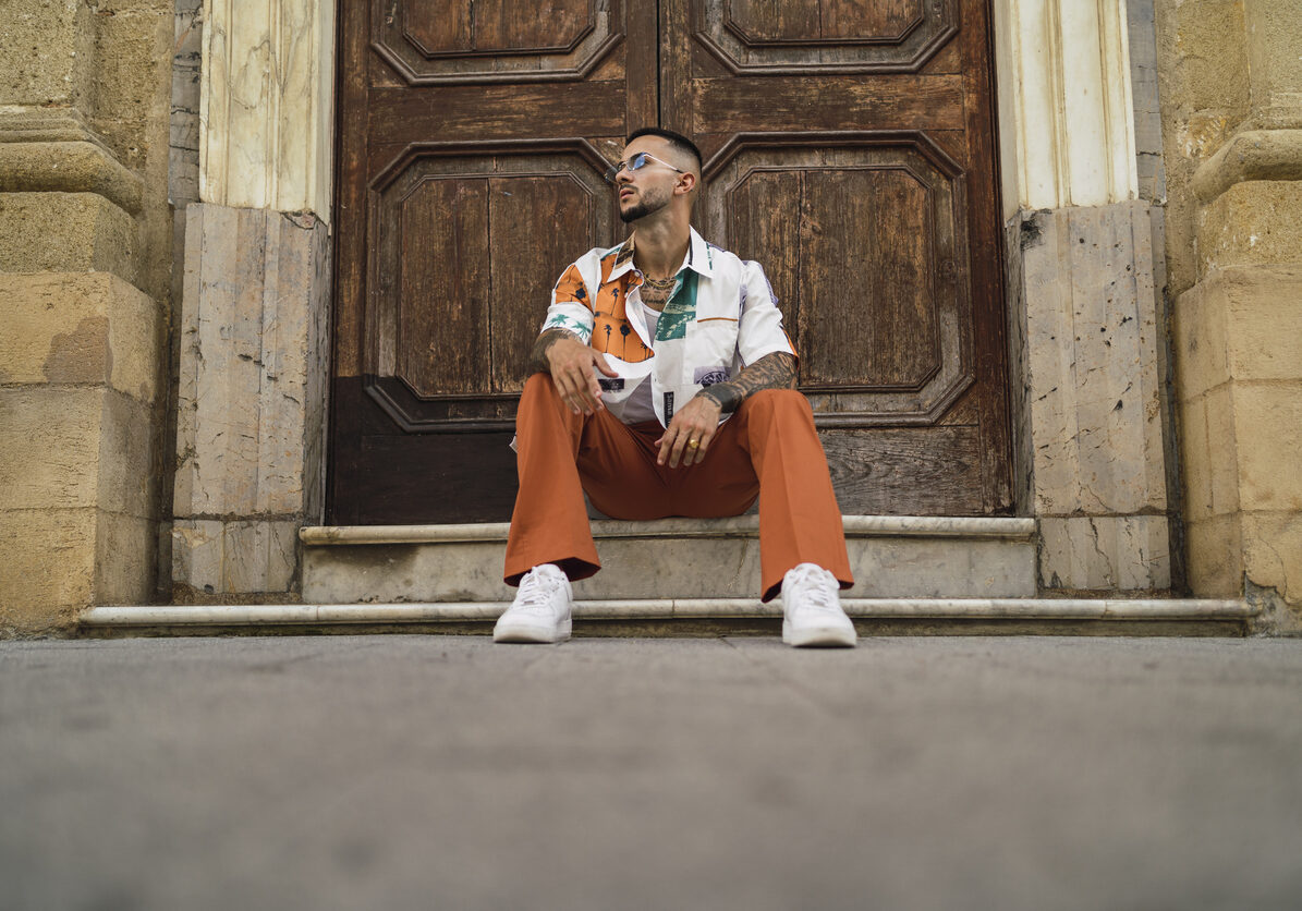 A hot caucasian man with arm tattoos with a patterned shirt and brown pants sitting on the entrance