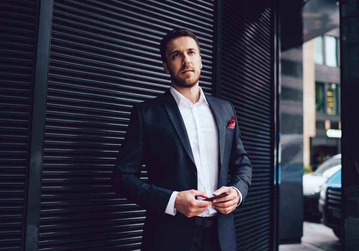 Confident smart businessman in dark suit with red scarf in pocket with mobile phone standing nearby metal wall and waiting looking away in downtown