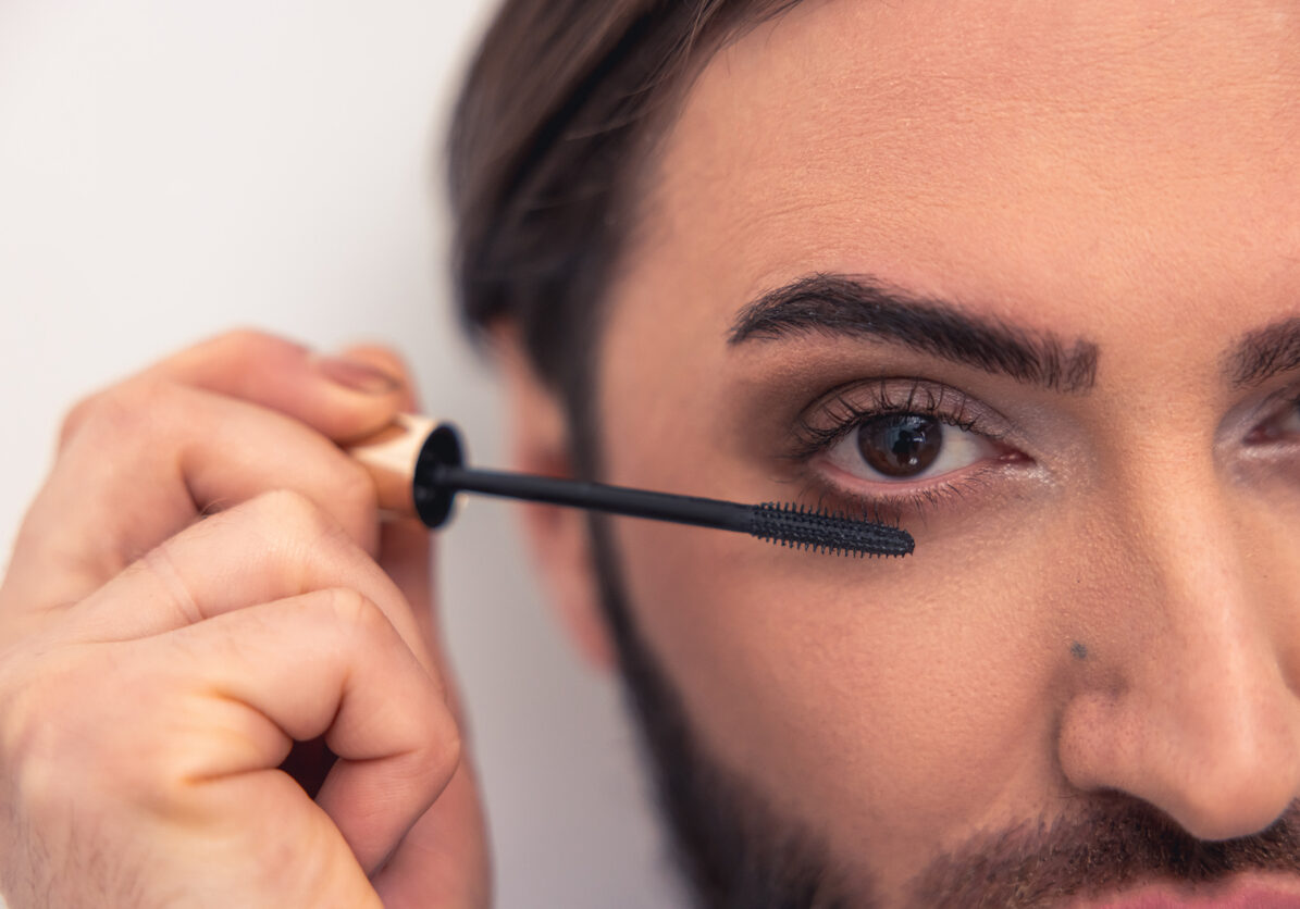 Cropped photo of a dark-haired man applying mascara to his long lashes using the wand