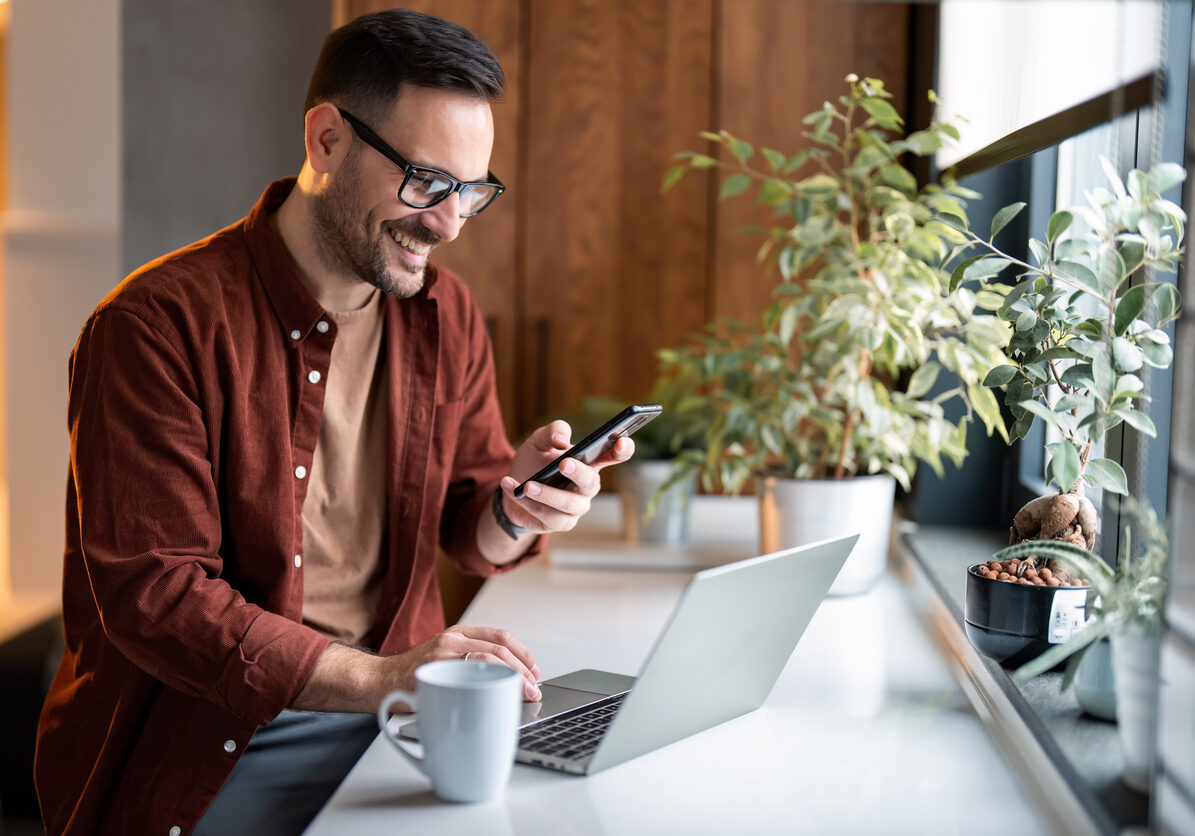 Satisfied modern millennial man in stylish casual clothes using smartphone and laptop computer for electronic banking, making reservation, online shopping and payments while spending time at home.