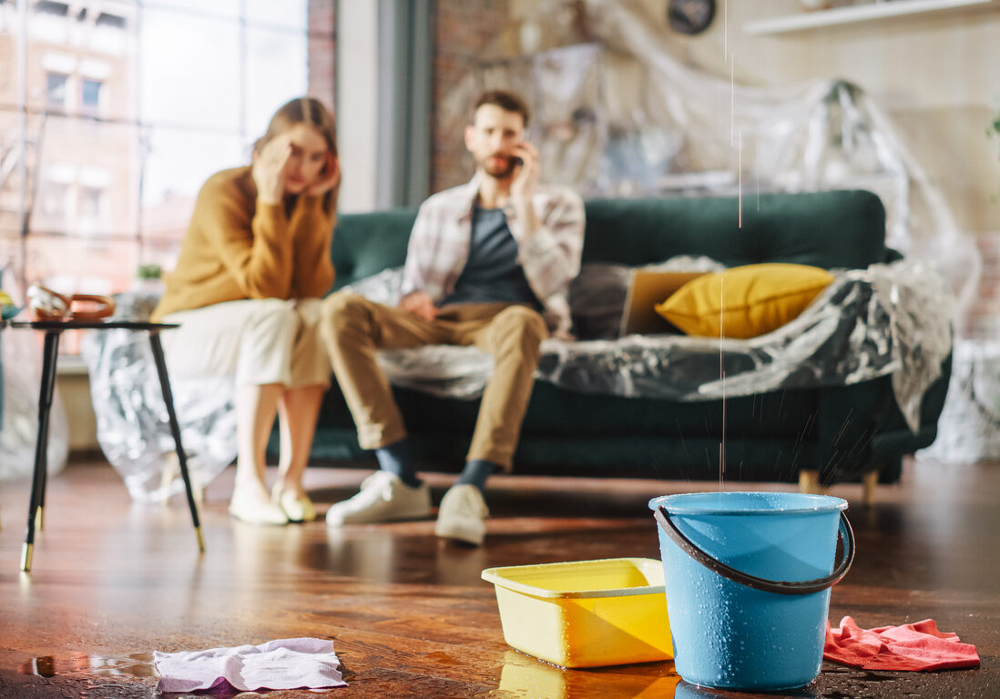 Roof is Leaking or Pipe Rupture at Home: Panicing Couple In Despair Sitting on a Sofa Watching How Water Drips into Buckets in their Living Room. Catastrophe, Distaster and Financial Ruin