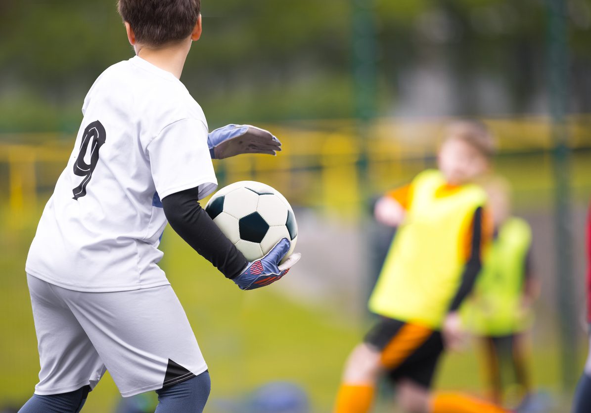 Young boy as a soccer goalie holding the ball in one hand ready to start a game. Football goalkeeper in jersey shirt and sports gloves throwing ball and playing a football match