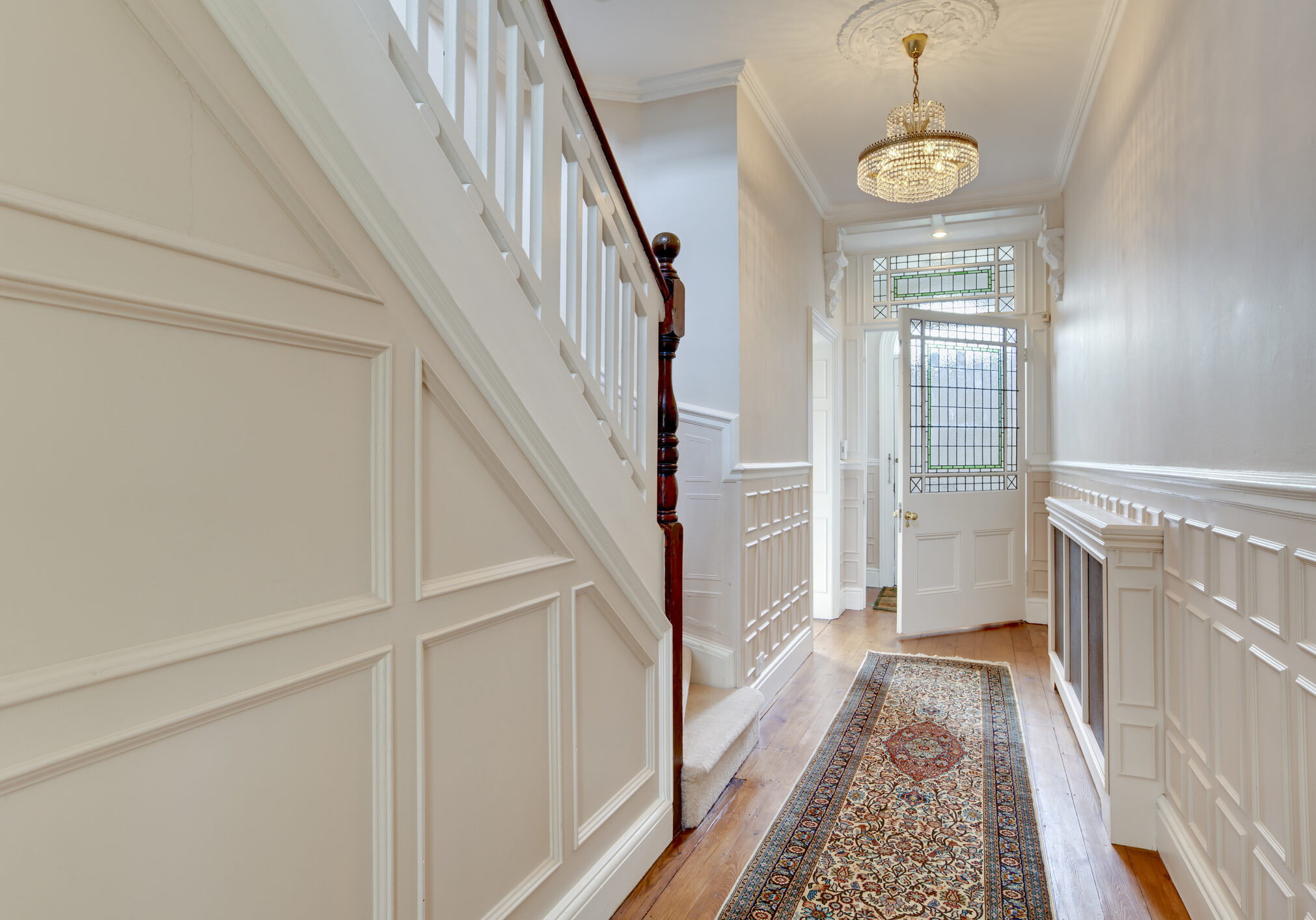 Reception hall to period victorian property decorated in white and cream with chandelier, stairs and glazed panelled door.