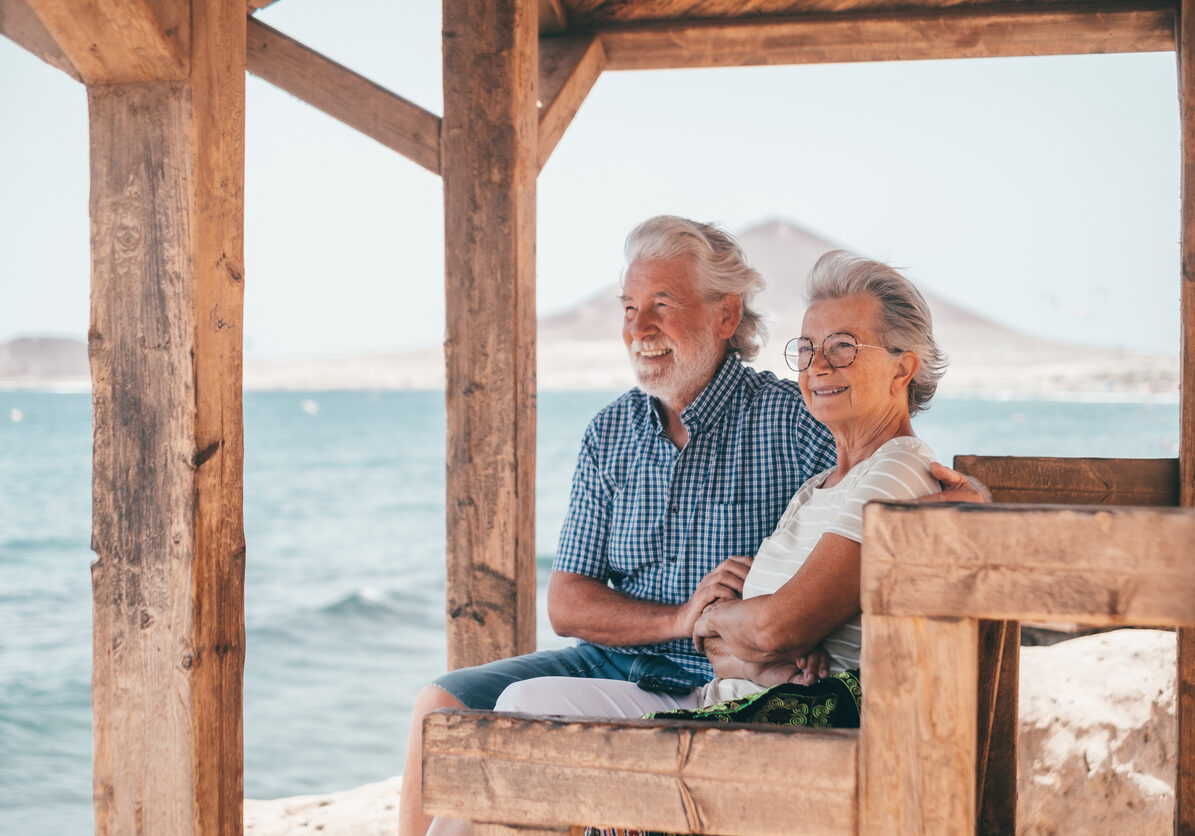 Lovely senior family couple sitting close to the beach looking at horizon over sea, two elderly people enjoying vacation or retirement together