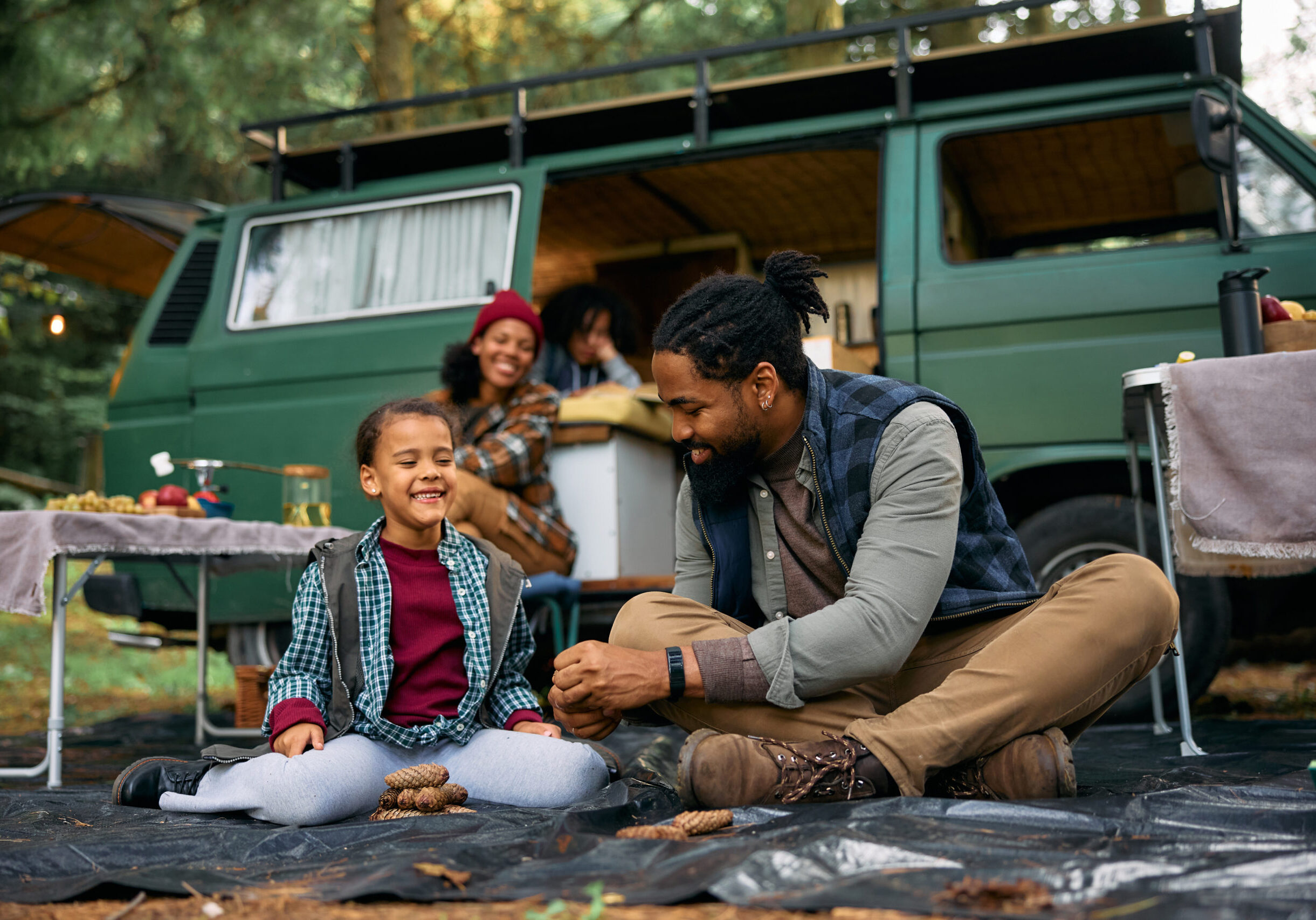 Happy African American little girl and her father having fun with pine cones by camper trailer in the woods.