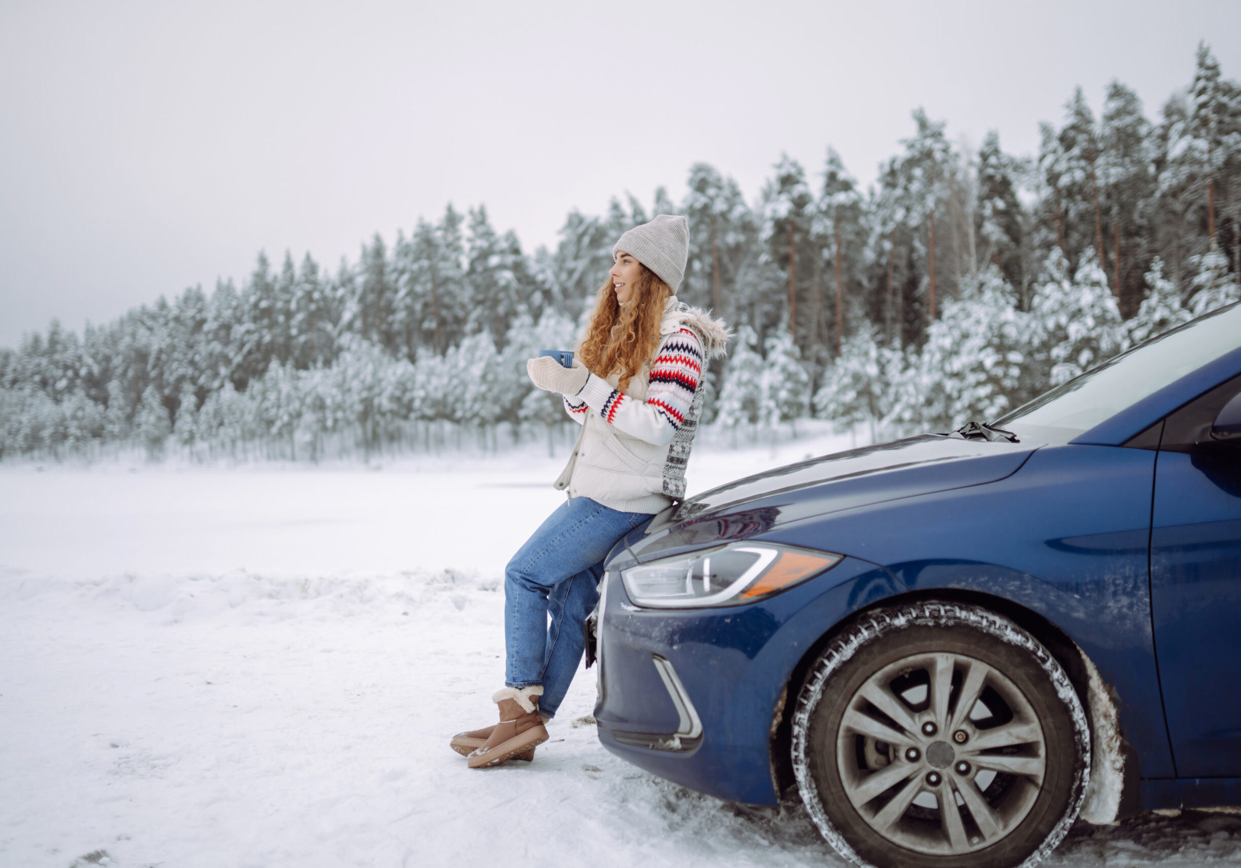 A smiling woman drinks a hot drink from a thermos while standing near her car on a winter snowy road in the forest. The concept of rest, freedom, relaxation, travel.