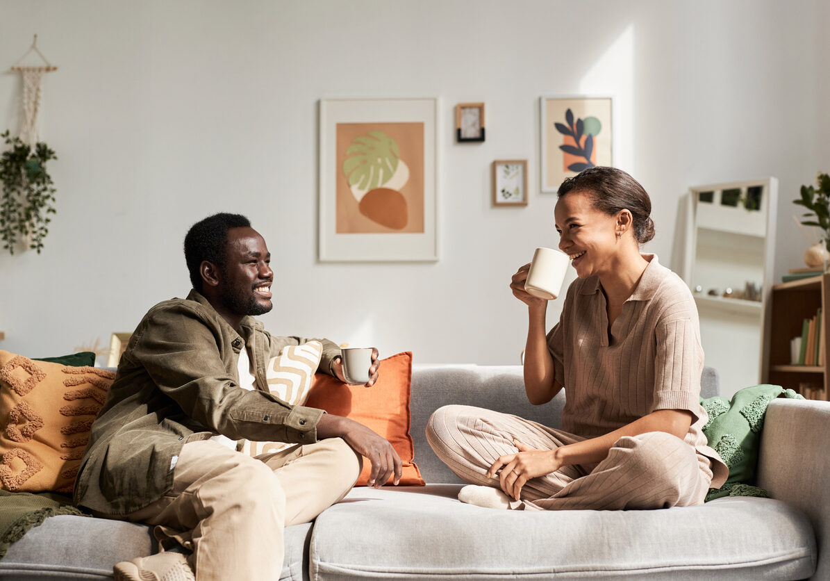 Side view portrait of young African American couple enjoying conversation sitting on comfortable couch in cozy home and drinking coffee